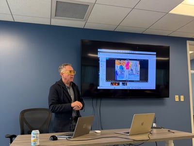 A man wearing yellow-tinted glasses stands in an office setting, presenting in front of a large screen displaying an image editing interface with two laptops on the desk in front of him.