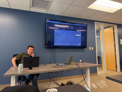 A man sits at a wooden desk in an office setting, looking thoughtfully towards the camera next to two laptops, with a large monitor displaying a software interface mounted on a blue wall behind him.