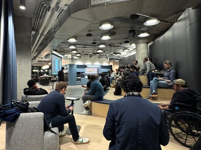 A group of people gathered in a modern, industrial-style indoor space, seemingly for a presentation or meeting. The room features tiered seating, exposed ceiling ducts, and a projection screen.