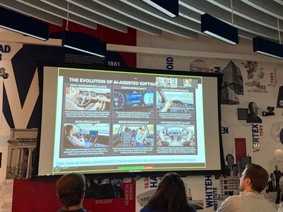 A photograph of a presentation about the 'Evolution of AI-Assisted Software' being shown on a large screen to an audience in a room with a detailed wall mural.