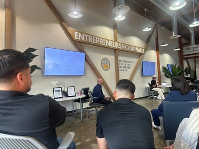 An indoor presentation or panel event taking place at the Entrepreneurs@Chapman space, featuring a speaker with a microphone and audience members watching from the foreground.