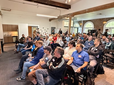 A man in a white t-shirt is giving a presentation to a large, seated audience in a modern office space with exposed brick walls and wooden beams.