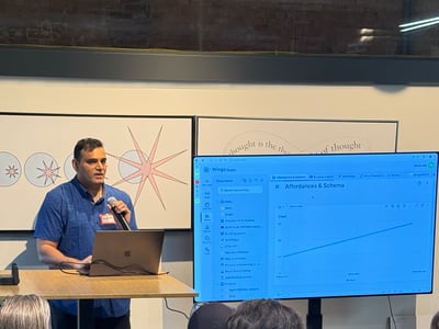 A man is giving a presentation to an audience, standing at a podium with a laptop and a large screen displaying a software interface.