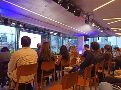 A group of people are seated in orange chairs, attending a presentation in a modern office space with large windows overlooking a city skyline at dusk.
