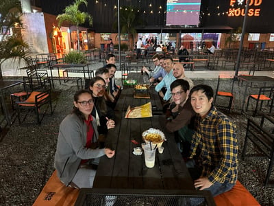A group of ten people are gathered around a long wooden table in an outdoor food court at night, smiling for a group photo.