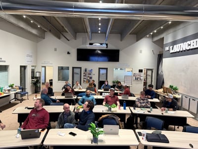 A candid photograph captures a group of approximately nineteen people, mostly men, seated at rows of tables in a modern, industrial-style conference room during a workshop or meeting, with many open laptops visible.