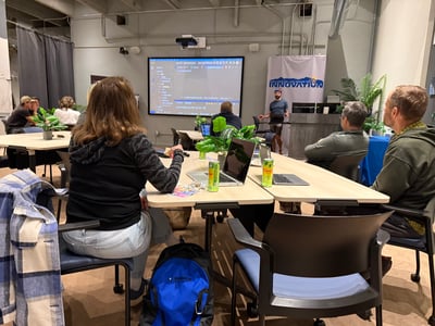 An indoor photograph showing a presentation session where a speaker stands near a large screen displaying code, with audience members seated at tables.