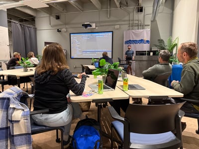A group of people sitting at tables in a classroom setting, watching a coding presentation displayed on a large screen.