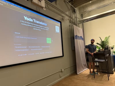 A man stands next to a large projection screen displaying a 'Yoda Translator' web application in an indoor presentation setting.