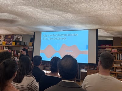 A photograph showing an audience from behind, watching a presentation slide projected on a screen in an indoor space filled with bookshelves.