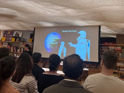 An indoor photograph showing an audience seated and watching a presentation on a large screen. The screen displays a variation of the 'Always Has Been' astronaut meme with the text 'wait, alignment is the bottleneck?' and 'always has been'.