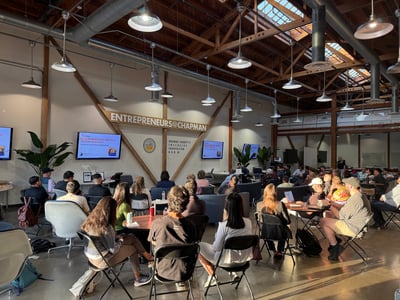 An indoor photograph of a presentation event at the Entrepreneurs@Chapman Innovation Hub, showing an audience seated in chairs facing screens and a presenter under a high wooden ceiling with exposed beams.