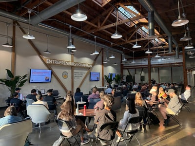 An indoor photograph of a presentation event at the Entrepreneurs@Chapman innovation hub, showing an audience seated in chairs and at small tables facing presentation screens under a high, wood-trussed ceiling.