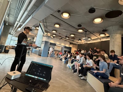 A man is delivering a presentation to a group of people seated on tiered benches in a modern, industrial-style office space.