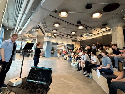 A woman gives a presentation to a seated audience in a modern, industrial-style office space with exposed concrete and piping. A man stands in the foreground near a laptop and monitor setup on a high table.