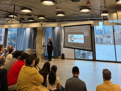 A presenter stands before a projector screen displaying 'VISION CLAW' while addressing an audience in a modern, industrial-style room.
