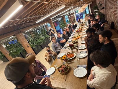 A group of people are gathered around a long wooden dining table inside a modern rustic event space, while a man in the foreground stands to speak to the attendees.