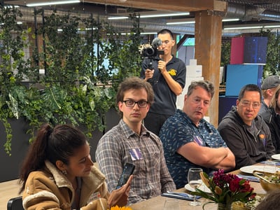 A group of people are gathered around a long wooden dining table filled with colorful dishes, salads, and floral centerpieces in a modern indoor setting.