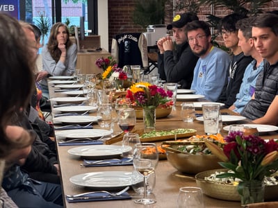 A group of people are seated along a long wooden dining table set with white plates, wine glasses, and vibrant floral centerpieces in an indoor setting.