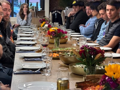 A group of people are seated along a long, elegantly set dining table decorated with floral centerpieces and filled with plates, glasses, and bowls of food.