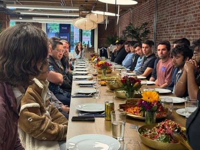 A large group of people are gathered around a long wooden dining table set with plates, food, and flowers in an indoor space with a brick wall.