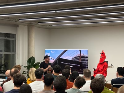 A man in a black t-shirt and cap is giving a presentation next to a person in a bright red lobster costume, in front of a seated audience.