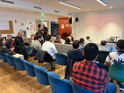 A group of people are seated in a modern office space, attending a presentation and looking towards a large projection screen.