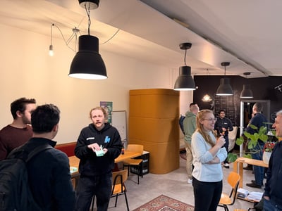 A candid photo showing a group of approximately nine people socializing indoors in a modern, casual space featuring black pendant lights and a mix of white and dark accent walls.