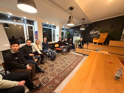 A photograph captures a small audience of people seated on chairs and a patterned rug, attending an informal evening event or presentation in a warmly lit indoor space with a stage area and a large wooden table in the foreground.