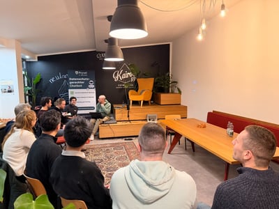 A group of people is gathered indoors, sitting on chairs and cushions, attending a casual presentation or panel discussion in a warmly lit event space. A small panel sits on a raised wooden platform against a black wall decorated with branding and a promotional banner.