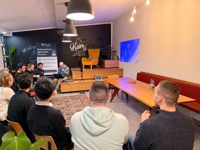 A group of people is gathered indoors, attending a presentation or discussion in a modern co-working space featuring a small stage, a chalkboard wall with branding, and a long wooden table. Several individuals are seated on the stage area, while the audience sits facing them, mostly captured from behind.