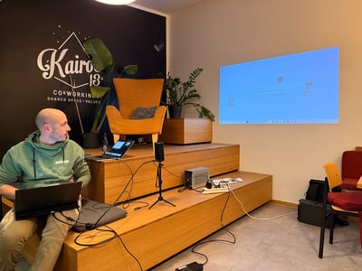 A man sits on tiered wooden seating in a co-working space, working on a laptop while looking toward a large, light-blue projected screen displaying a graphical interface.