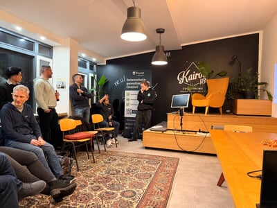 A candid photograph capturing an informal evening event or presentation held in a co-working space, featuring a group of people gathered around a small stage area with a distinctive orange armchair and branded black wall.
