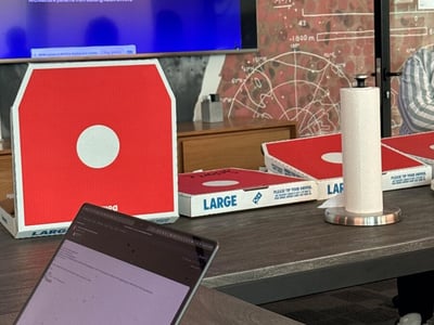 A photograph of an office meeting room featuring several pizza boxes on a table, a laptop in the foreground, and a large screen displaying a presentation about autonomous coding agents.