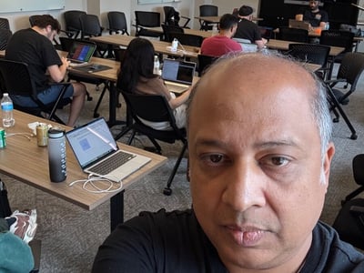 A man takes a selfie in a classroom or office setting where several other people are working on laptops at long tables.