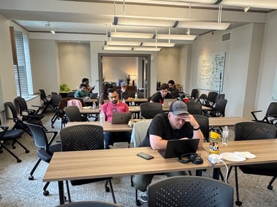 A group of people are seated at long tables in a modern indoor workspace, focused on their laptops.