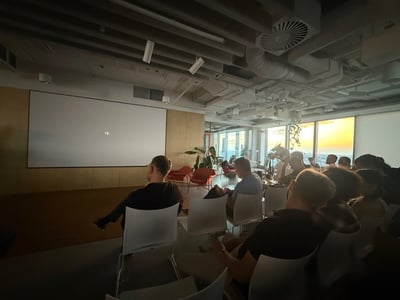 An audience sits in rows of chairs inside a modern office presentation room, facing a large blank projection screen with a sunset visible through the windows on the right.