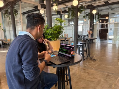 A man and a woman are sitting at a high table in a modern office space, focused on a laptop screen displaying lines of code.
