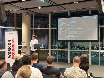 A man stands at a podium giving a technical presentation, with a large screen behind him displaying Python code to an audience.