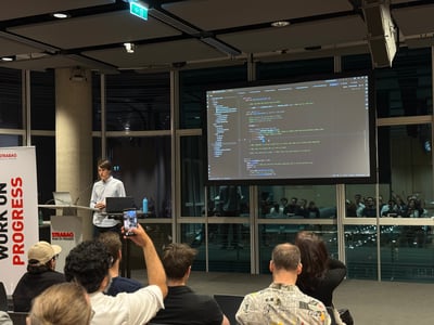 A man stands at a podium giving a technical presentation, with a large screen behind him displaying Python code to an audience.