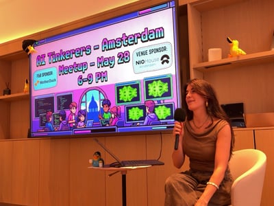 A woman sits holding a microphone next to a large digital display advertising an AI Tinkerers meetup in Amsterdam.