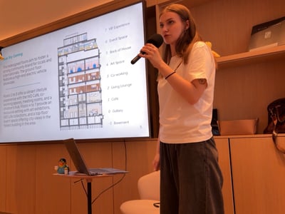 A young woman stands in an indoor setting, holding a microphone and presenting in front of a large screen displaying an architectural cross-section diagram of a building.