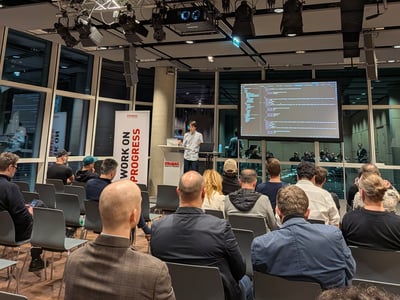 A man stands at a podium giving a presentation to an audience in a modern indoor space, with a screen displaying code and a banner that reads 'WORK ON PROGRESS'.