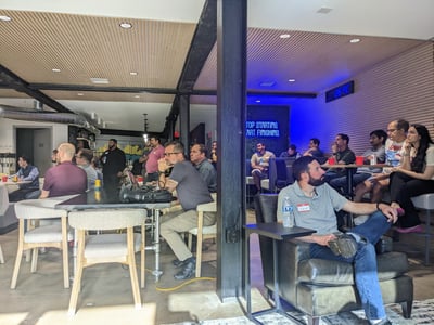 A group of people are gathered in a modern, industrial-style indoor space, seemingly attending a presentation or social event, with most looking towards the left side of the frame.