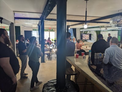A group of people are gathered in a modern office or coworking space, watching a presentation displayed on a monitor.