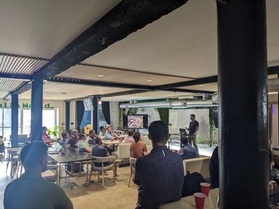 A group of people are gathered in a modern office or event space watching a presentation given by a man standing near a screen and whiteboard.