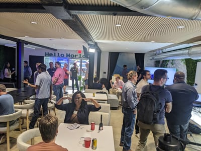 A group of people gathered in a modern, industrial-style indoor space for a social or networking event, with some sitting at tables and others standing in conversation.