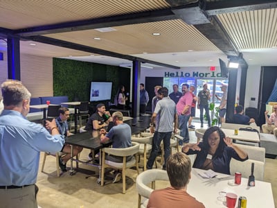 A group of people are seen networking and socializing in a modern, open-plan office or event space featuring a 'Hello World' sign and a green living wall.