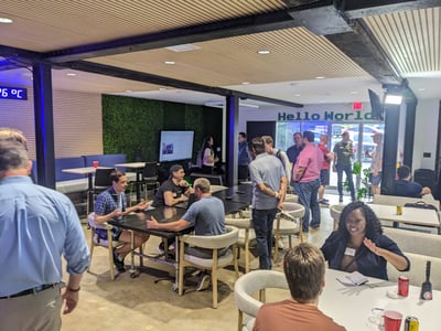 A group of people are gathered in a modern, open-plan indoor space with wood-paneled ceilings and a green plant wall, appearing to be at a networking event or social gathering.