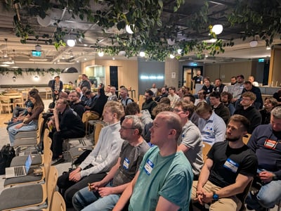 A large group of people, mostly men, are seated in a modern indoor event space attending a presentation or meetup.